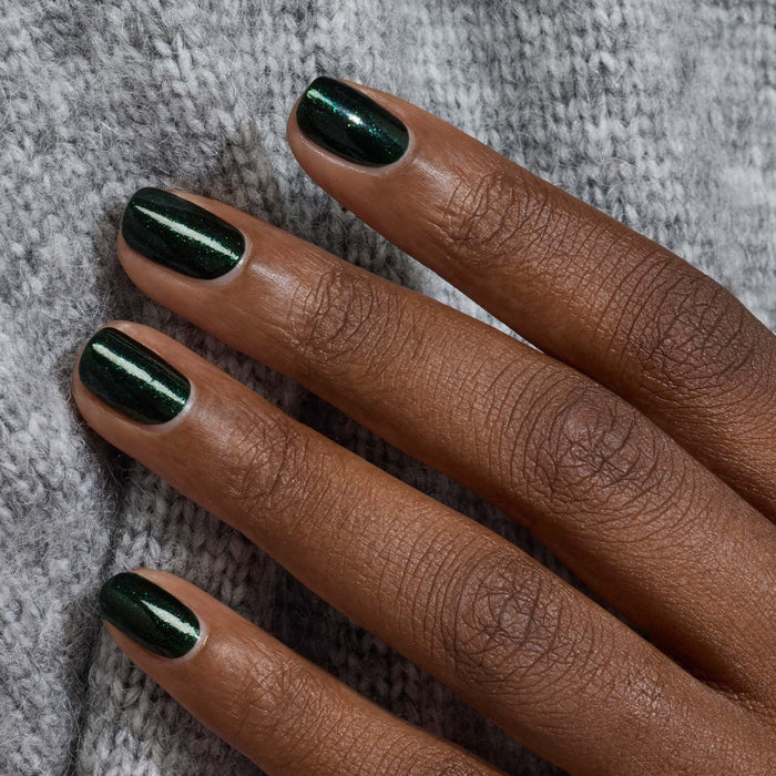 A close-up of a hand with glossy Manucurist Poison gel polish in forest green, resting on light gray knit fabric.