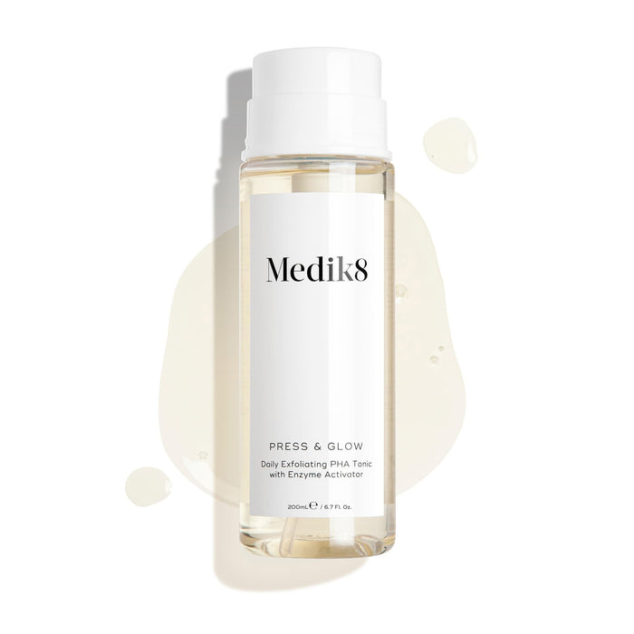 Medik8 Press & Glow tonic bottle stands against a clean white background with translucent liquid droplets and a puddle, highlighting its watery texture. Text identifies the product as a 200mL "Daily Exfoliating PHA Tonic with Enzyme Activator."