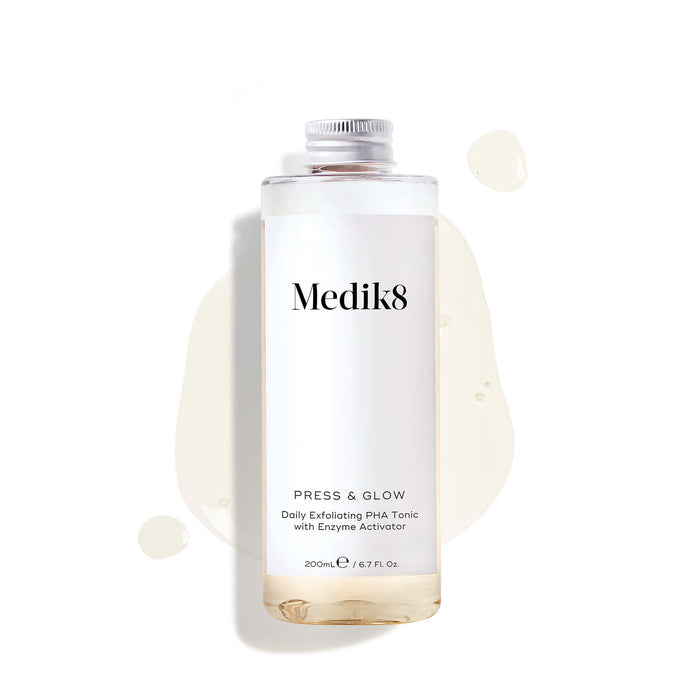 A 200mL clear refill bottle of Medik8 Press & Glow exfoliating tonic featuring a silver screw cap. Set against a white background, the bottle sits amidst a puddle and droplets of the pale yellow tonic, showcasing its liquid texture. The label text reads "Daily Exfoliating PHA Tonic with Enzyme Activator."