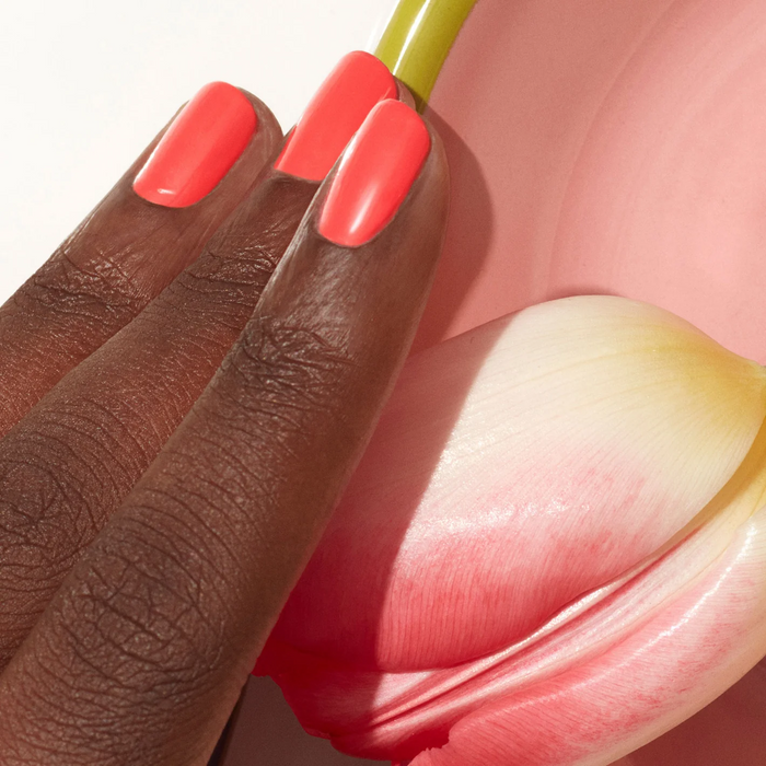 A hand with dark skin and neatly manicured nails painted in Manucurist’s Pulp coral pink polish rests on a pink and white tulip beside a pink dish with a yellow rim.