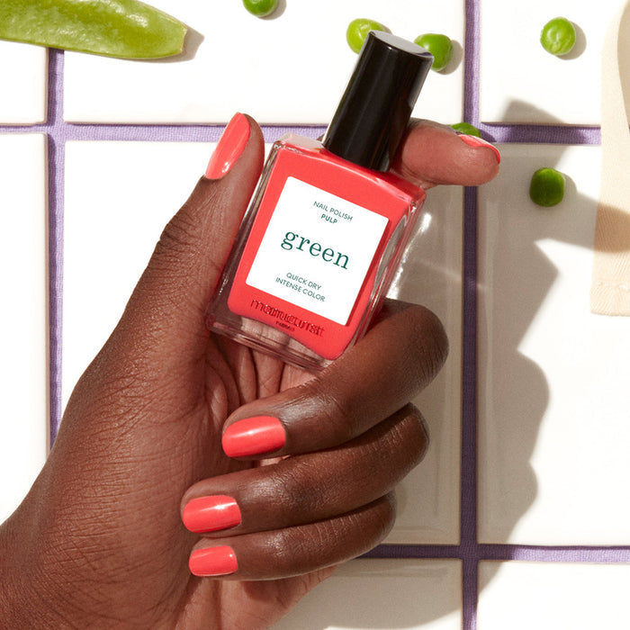 A hand with medium-dark skin tone displays neat coral nails while holding a Manucurist Pulp nail polish bottle. The background has green peas and a white tiled surface with lavender grout.