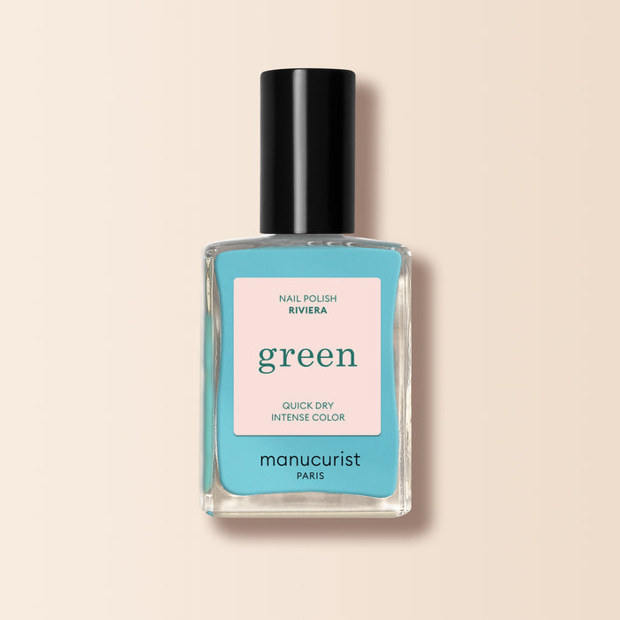 A bottle of Manucurist Riviera nail polish from the Green™ range has a black cap, beige-pink front label, and contains energizing blue polish. The background is soft light beige.