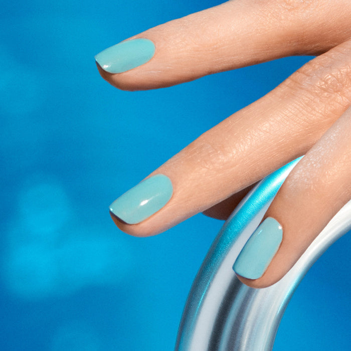 A close-up of a hand with Manucurist's Riviera glossy turquoise blue nail polish gripping a shiny metal pool ladder, with an energizing blue pool in the background.