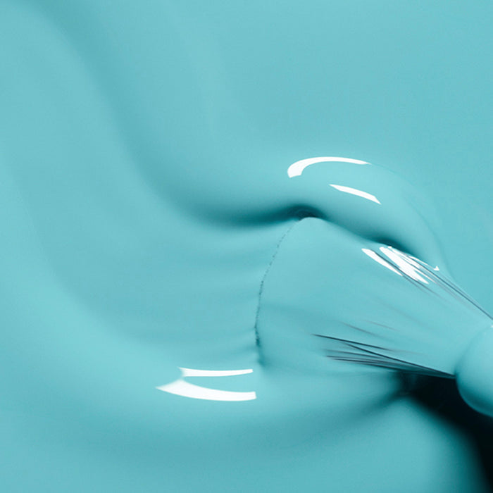 A close-up of a paintbrush applying Manucurist's Riviera, a glossy turquoise blue nail polish, with visible bristles creating gentle waves in the wet color.