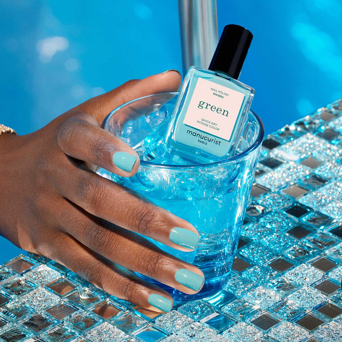 A hand with turquoise blue Riviera by Manucurist nail polish holds a glass of blue iced drink, with the matching nail polish bottle on the ice, set beside a sparkling pool.