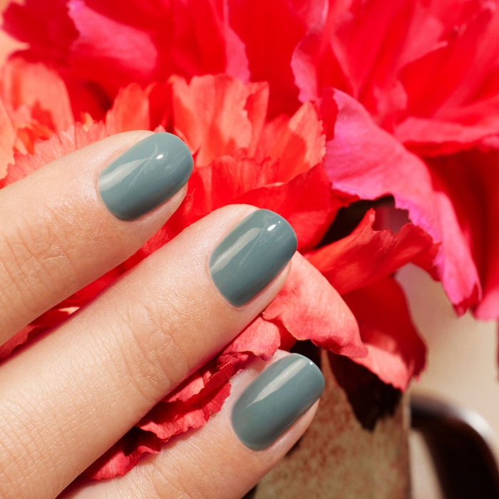 A close-up of a hand with short, glossy nails painted in "Sage" by Manucurist, held before vibrant pink and red carnation flowers.
