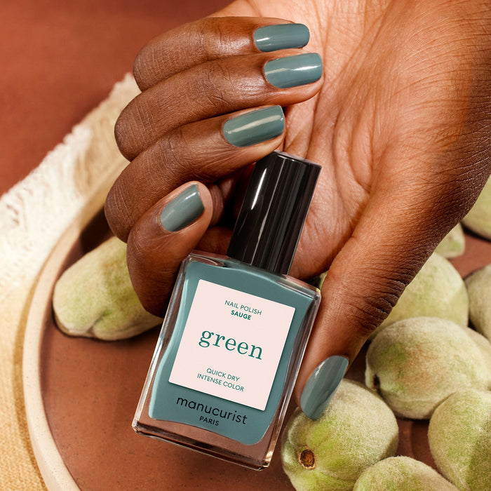 A hand with neatly manicured nails painted in "Sage" green nail polish holds a matching Manucurist bottle, with fuzzy green almonds in the background.