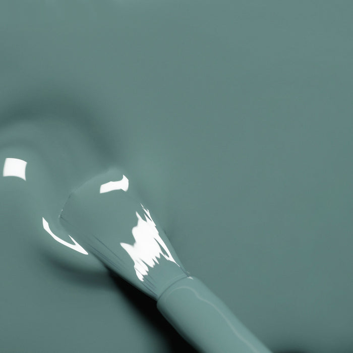A close-up of a Manucurist Sage nail polish brush coated in glossy, muted sage green polish rests on a matching liquid surface with reflected highlights.