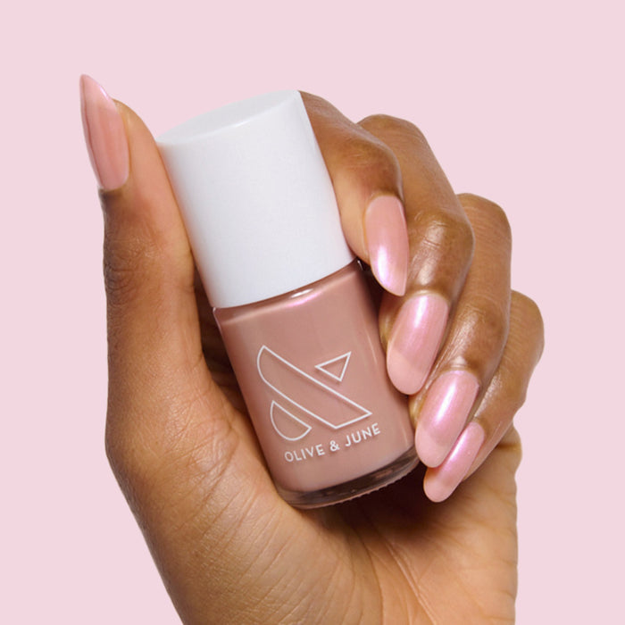 Hand with almond-shaped nails painted in iridescent Sandy Scallop gel polish holding Olive and June nail polish bottle