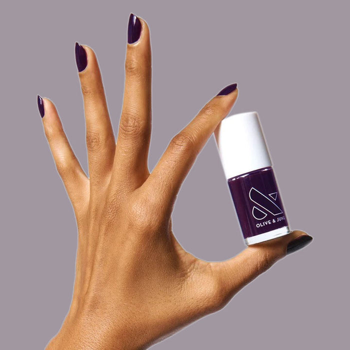 Hand with nails painted in deep plum shade holding SD nail polish bottle against gray background
