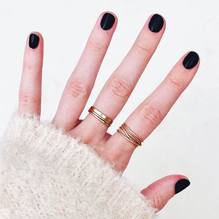 Hand with nails painted in deep black shade using SD nail polish, styled with gold rings and cozy sweater sleeve visible