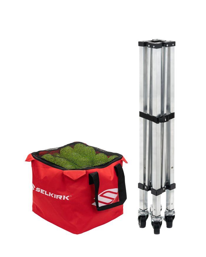 SELKIRK red canvas ball bag filled with green pickleballs beside folded aluminum net frame with caster wheels