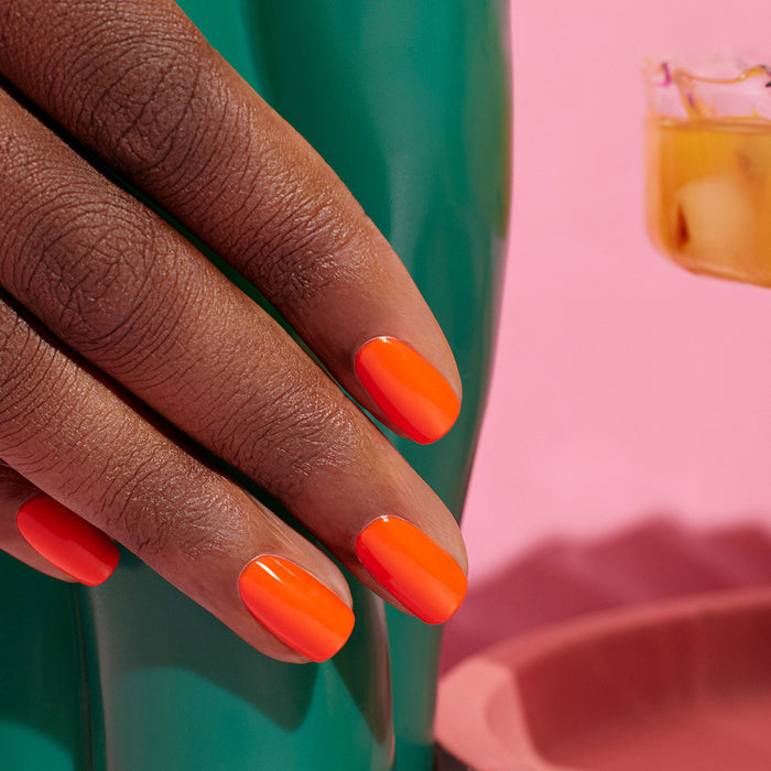 A close-up of a hand with smooth, medium brown skin and neatly manicured nails painted in Manucurist’s Green™ nail polish in the shade Sunset, resting on a glossy green surface, with a pink background and a glass of light drink.