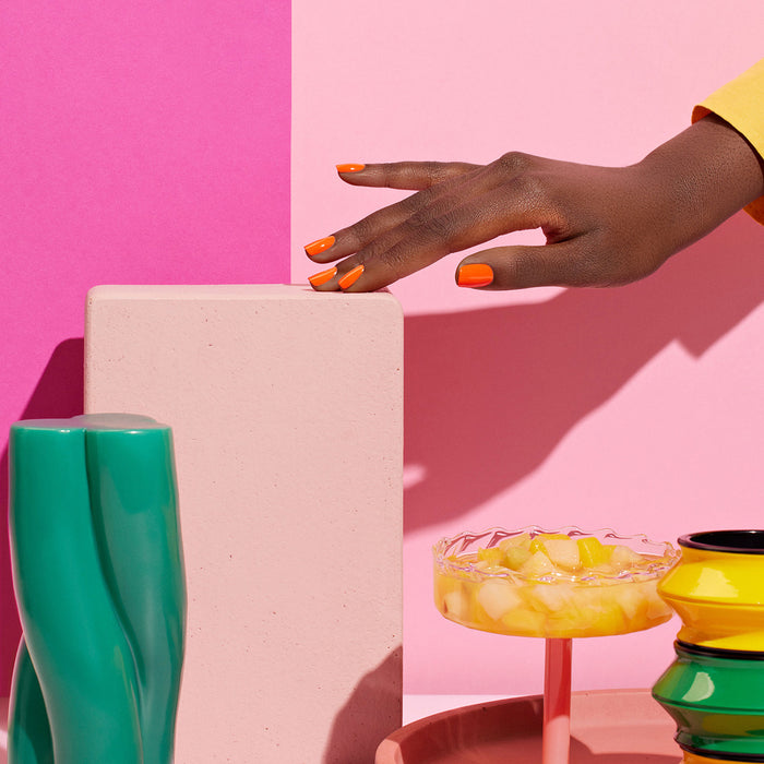 A hand with Manucurist's Sunset orange nail polish touches a pale block, surrounded by lively items like a Green™ range vase, a yellow dessert in a glass dish, and a striped vase, all set against a pink background.