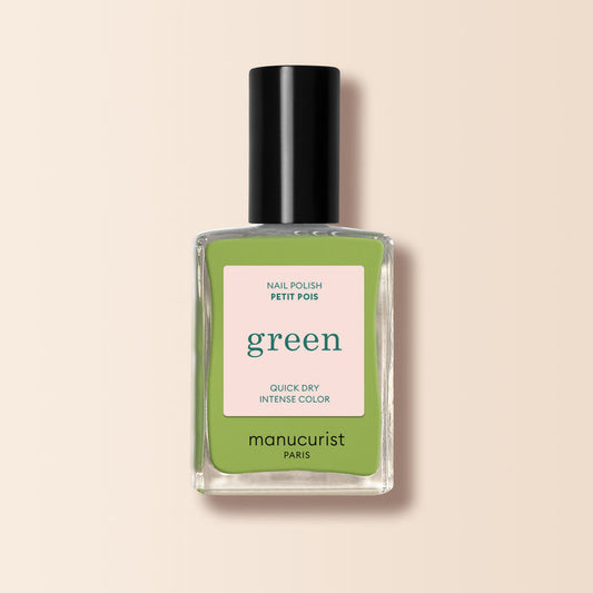 A bottle of Manucurist Green™ nail polish with a black cap, light green liquid, and a label reading Green, Quick Dry Intense Color, Nail Polish Sweet Pea. The background is light beige.