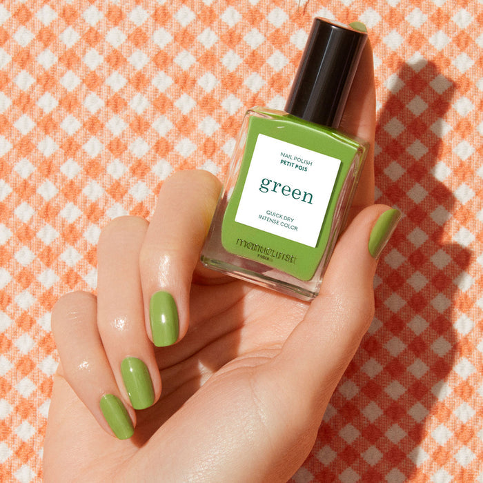A hand with green painted nails holds a bottle of Manucurist Sweet Pea nail polish against an orange and white checkered fabric background.