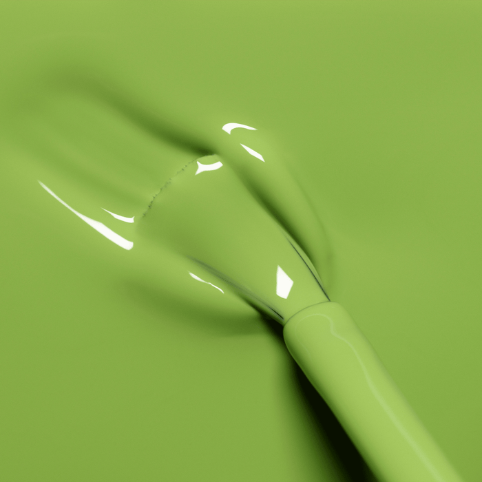 A close-up of a nail polish brush spreading glossy, light green Sweet Pea by Manucurist on a smooth surface. The thick, shiny polish reflects light softly.