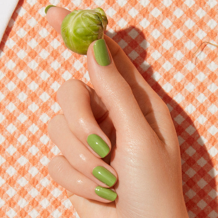 A hand with Manucurist's Sweet Pea green nail polish holds a Brussels sprout against an orange and white checkered fabric background.