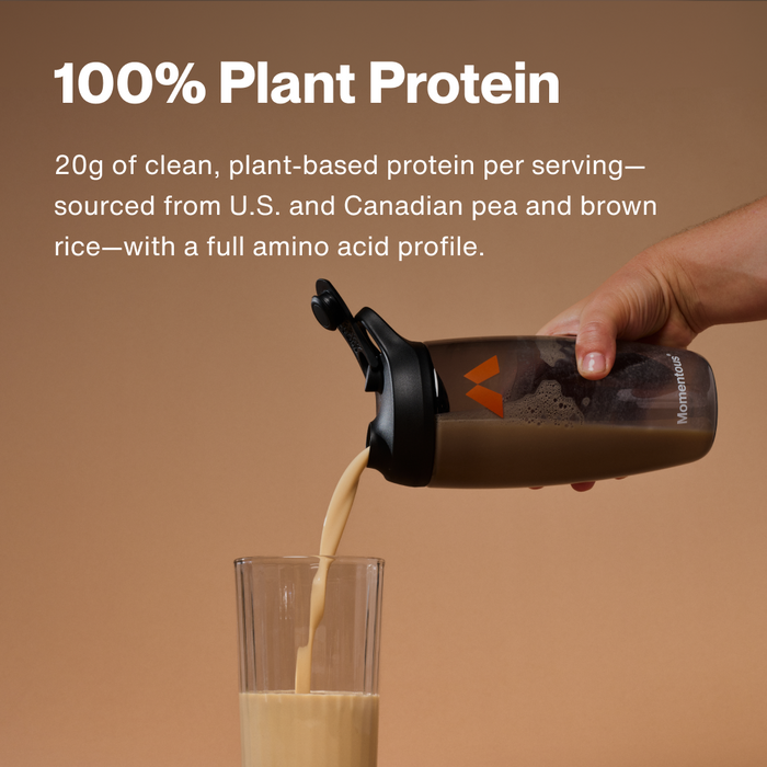 Hand pouring tan-colored protein powder from black shaker bottle into glass against beige background.
