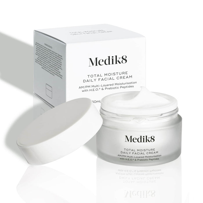 An open frosted glass jar of Medik8 Total Moisture Daily Facial Cream with a white lid resting beside it, displaying its rich white cream. The jar stands in front of its white cardboard carton on a light background. Text on both reads: "AM/PM Multi-Layered Moisturisation with H.E.O.® & Prebiotic Peptides."