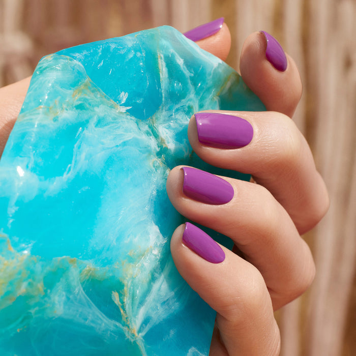 A hand with neatly manicured nails painted in Violette by Manucurist holds a large, rough turquoise-colored stone. The background features softly blurred warm brown tones.