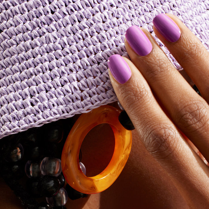A hand with smooth Violette nail polish by Manucurist touches a lavender woven hat. Chunky orange earrings and black beaded accessories add stylish flair in the background.