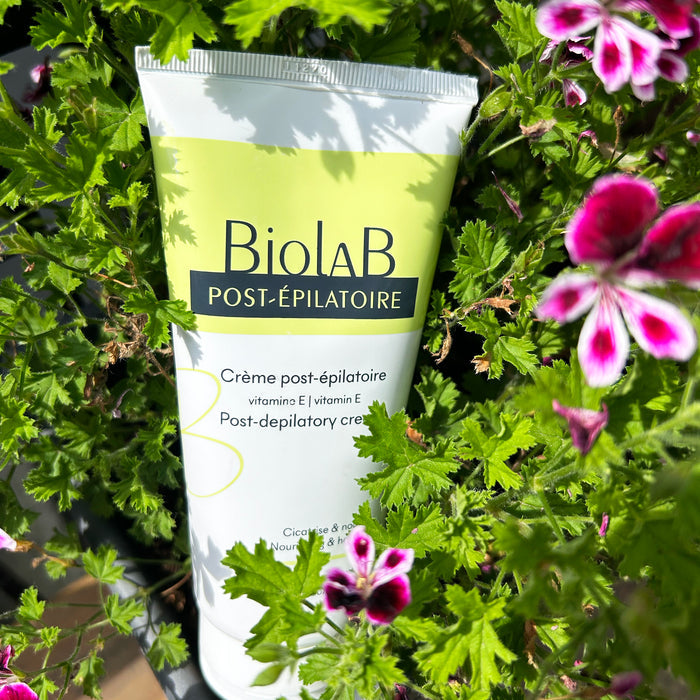 BiolaB Post-Épilatoire cream with vitamin E, nestled among vibrant green leaves and pink flowers for soothing skincare.