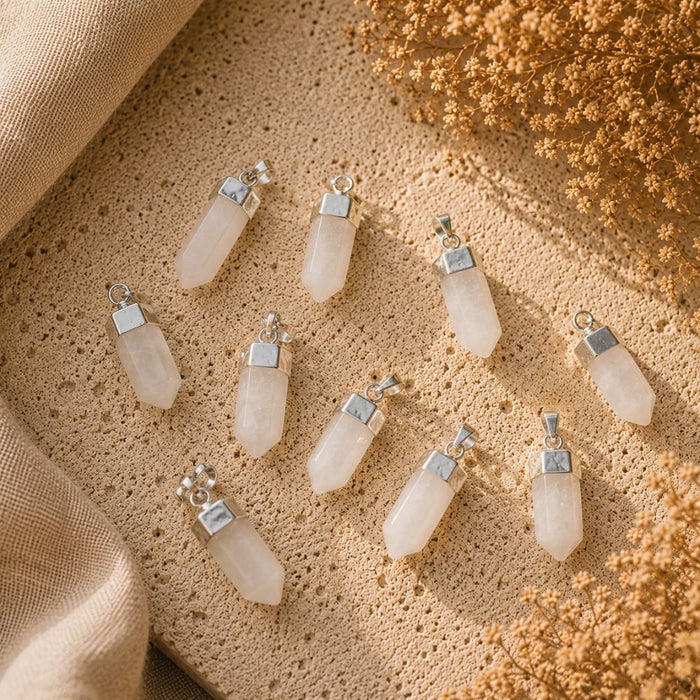 Nine pale pinkish-white crystal point pendants with silver caps are arranged on a textured beige stone surface with draped fabric and dried flowers.