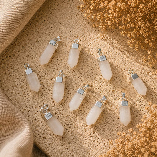 Nine pale pinkish-white crystal point pendants with silver caps are arranged on a textured beige stone surface with draped fabric and dried flowers.