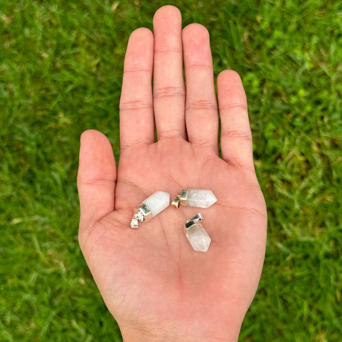 Four small white crystal point charms with silver metal caps resting on an open hand, set against a blurred green grass background.