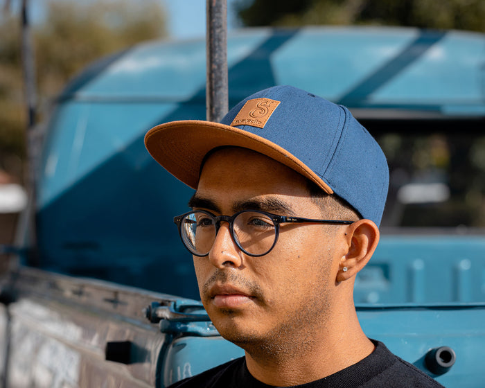 man wearing - blue suavecito hat with brown bill, brown text and brown background "S suavecito"